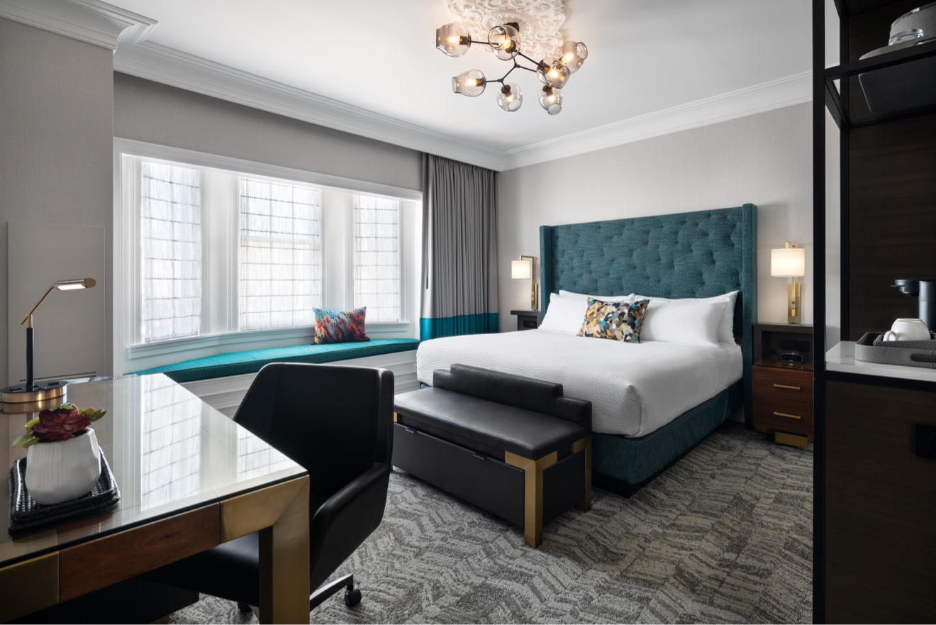 Guest room with teal upholstered bed, work desk, and city windows