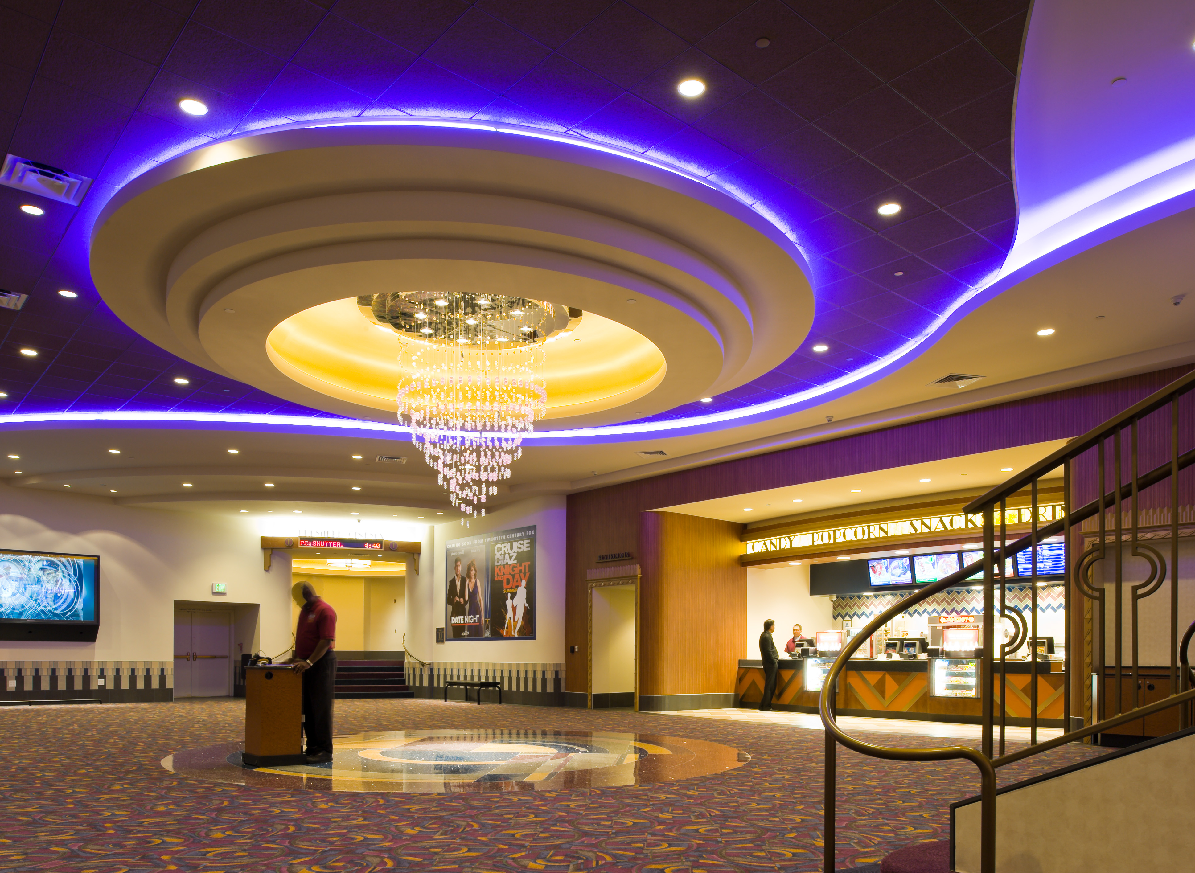 Cinema lobby with a circular tiered ceiling, purple cove lighting, and a crystal chandelier
